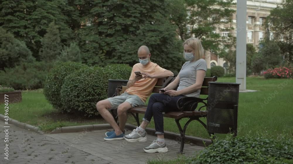 A man and a woman wearing protective masks sit at opposite ends of a bench and use their phones in the park A man and a woman wearing protective masks sit at opposite ends of a bench and use their phones in the park