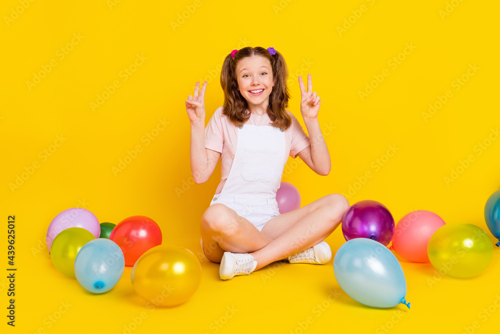 Fototapeta premium Portrait of pretty cheerful girl sitting on floor with air balls decor showing v-sign isolated over bright yellow color background