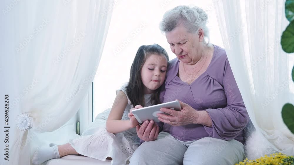 Cute small preschool little girl teaching old mature grandma nanny using digital tablet. Two generation family grandparent and grandchild exploring new technologies pad device tech at home Cute small preschool little girl teaching old mature grandma nanny using digital tablet. Two generation family grandparent and grandchild exploring new technologies pad device tech at home