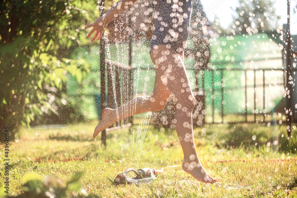 Small girl playfully jumping through sprinkler and water - obrazy, fototapety, plakaty 