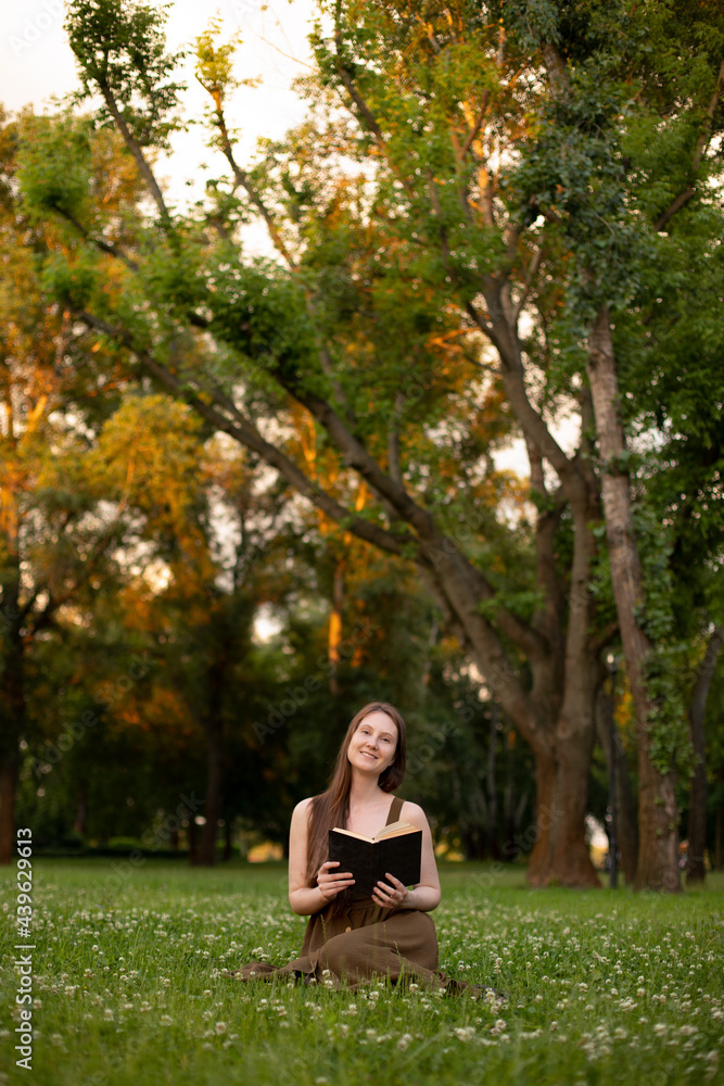 girl in an olive dress reads a book while sitting on the green grass in the park. place for your design. the summer vacation concept