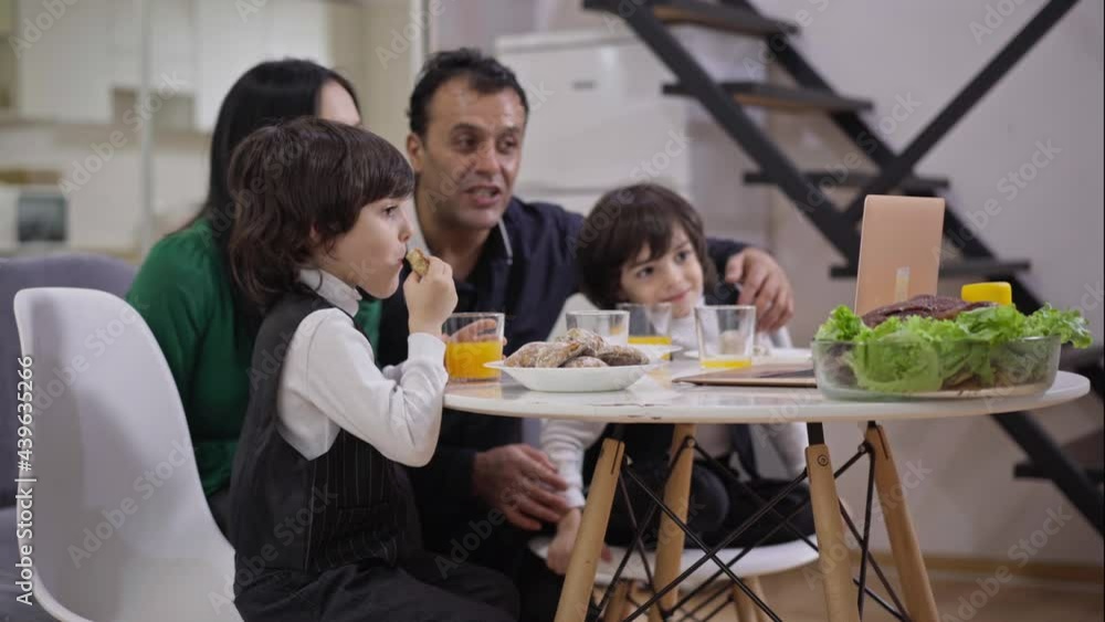 Side view of absorbed little boy eating sweet tasty bakery with blurred joyful mother father and twin brother laughing watching comedy movie on laptop at background. Happy family dinner indoors Side view of absorbed little boy eating sweet tasty bakery with blurred joyful mother father and twin brother laughing watching comedy movie on laptop at background. Happy family dinner indoors