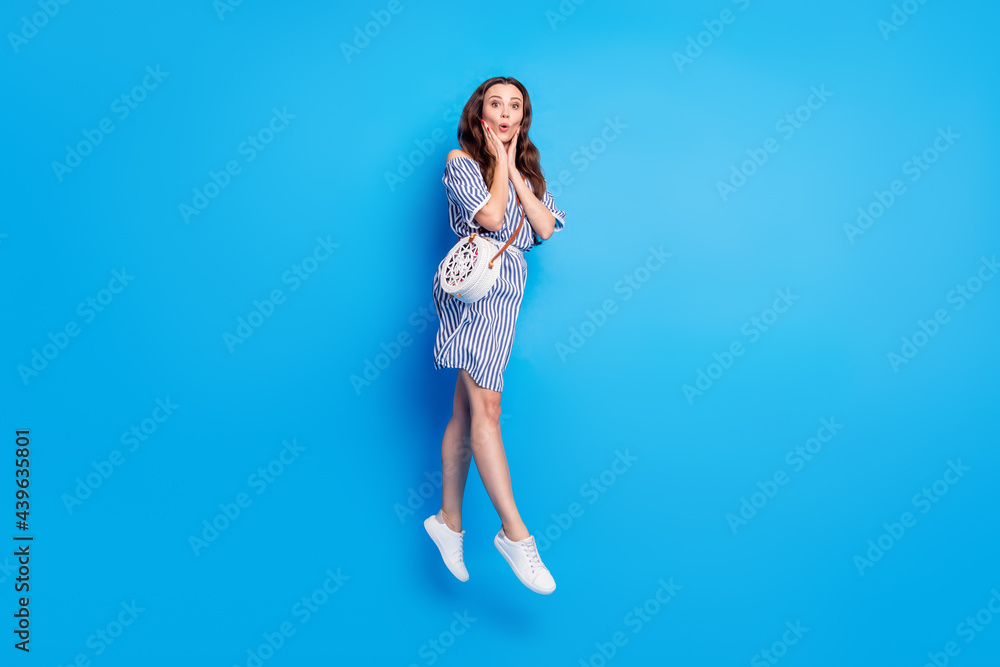 Photo of impressed cute young woman dressed striped outfit handbag arms cheeks jumping high isolated blue color background Photo of impressed cute young woman dressed striped outfit handbag arms cheeks jumping high isolated blue color background