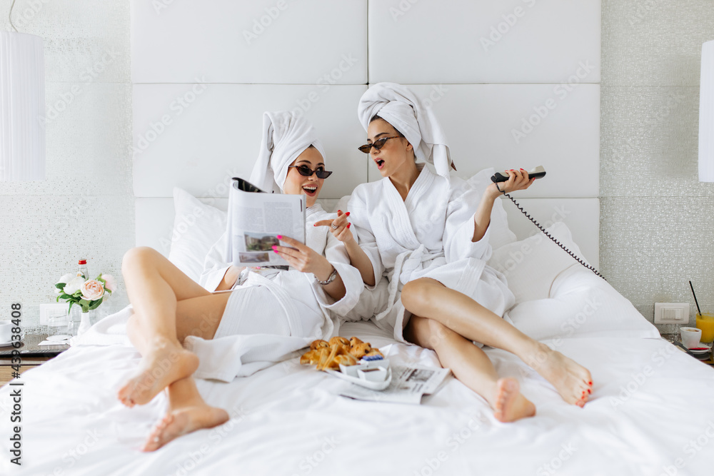 Two beautiful woman in bathrobe reading magazine, having fun and calling room service