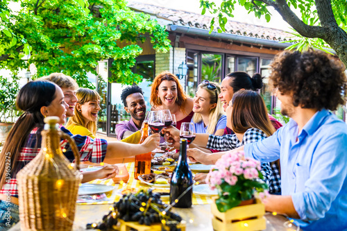 Happy multi ethnic friends having fun outdoor toasting red wine glass at winery on sunset - Young people enjoying time together at table in the restaurant garden © charmedlightph