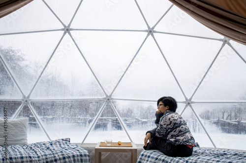 Thoughtful woman in spherical camp cupola in winter forest sitting inside of a  transparent tent with a phone 