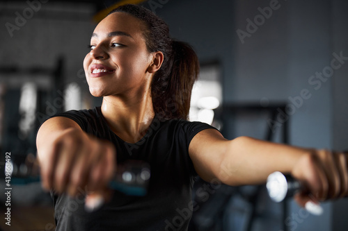 Cheerful pretty woman exercising shoulders in gym