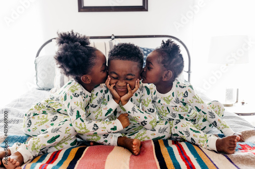 Three siblings in matching pajamas sitting on the bed. The two sisters are kissing their brother.  photo