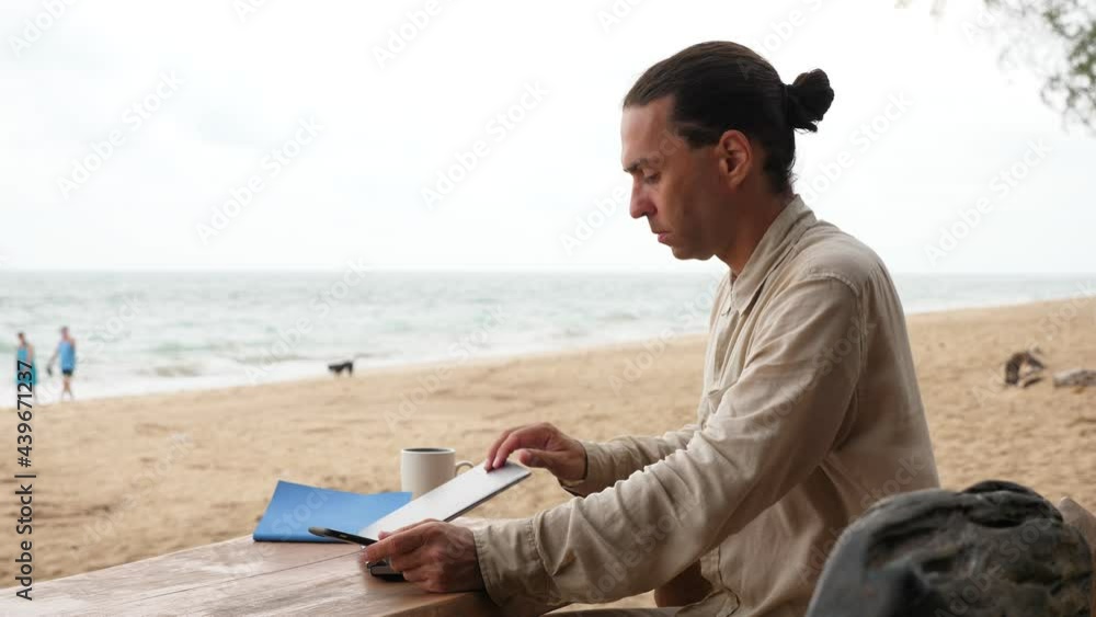 Guy shut down laptop and start using phone. Freelancer sit at wooden table, outdoor working place against sea beach. Portrait shot of man, blurred background. Digital nomad concept Guy shut down laptop and start using phone. Freelancer sit at wooden table, outdoor working place against sea beach. Portrait shot of man, blurred background. Digital nomad concept