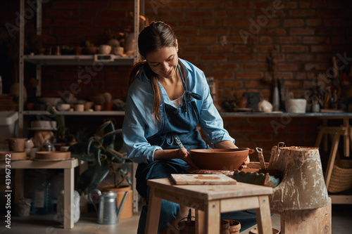Friendly attractive female artisan wearing apron modeling ceramic tableware in pottery workshop