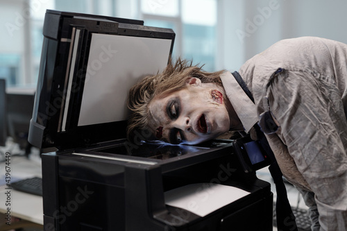 Gloomy man with zombie greesepaint on face keeping head on screen of xerox machine photo