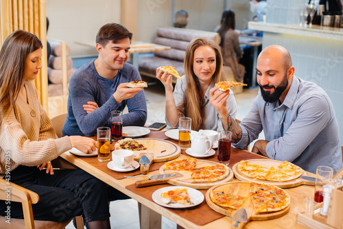 A group of young cheerful friends is sitting in a cafe talking and eating pizza. Lunch at the pizzeria.