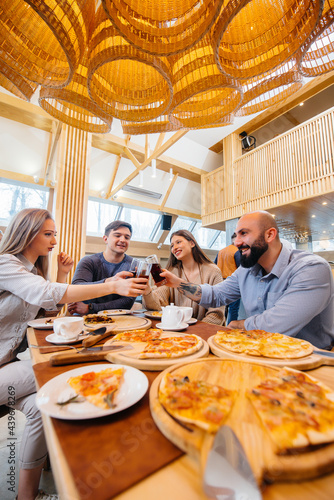 A group of young cheerful friends is sitting in a cafe talking and eating pizza. Lunch at the pizzeria.