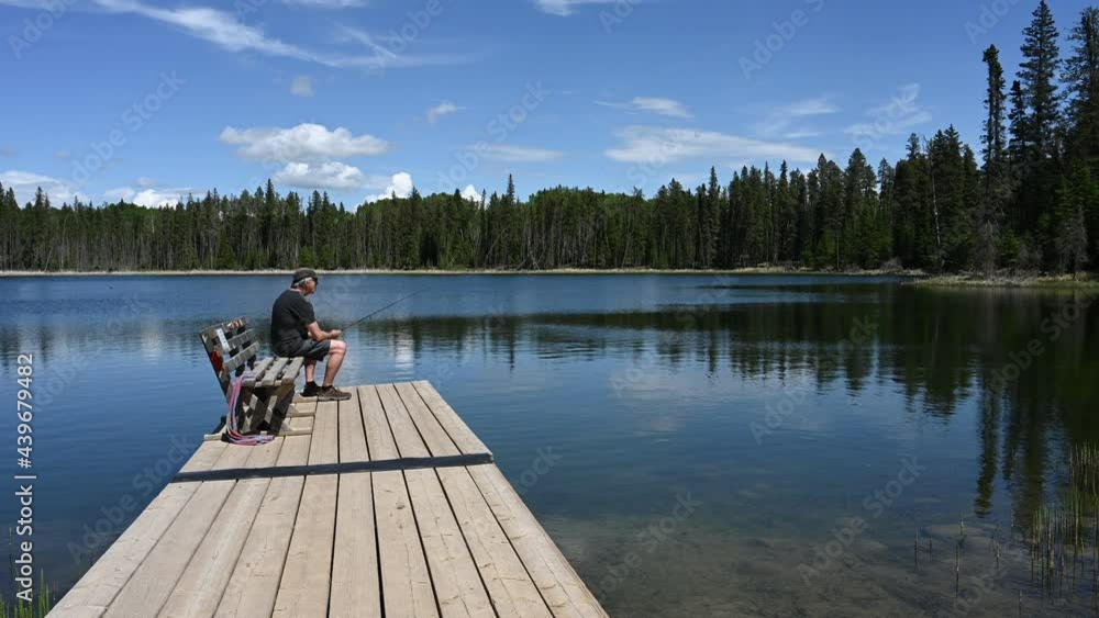 Gray haired man in shorts sitting on a bench on a wooden dock is fishing in a small lake.
Gray haired man in shorts sitting on a bench on a wooden dock is fishing in a small lake.