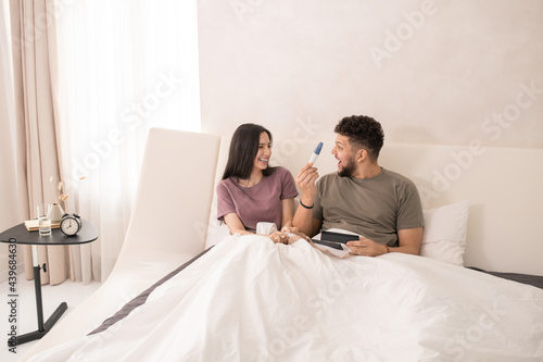 Happy woman looking at her surpwised husband with pregnancy test while lying in bed