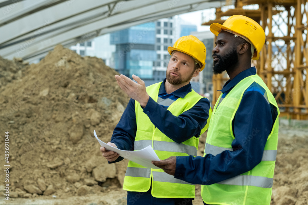 Two young intercultural builders in uniform discussing sketch and new construction Two young intercultural builders in uniform discussing sketch and new construction