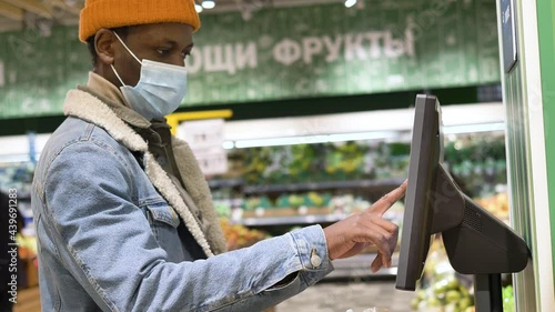 Black man in blue disposable face mask and denim jacket weighs oranges in transparent plastic bag on grey self-checkout close view photo