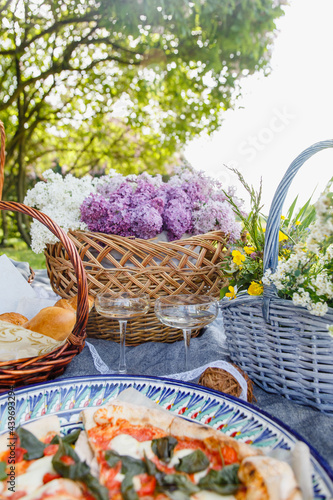 Fototapeta Naklejka Na Ścianę i Meble -  
Wicker basket with lilacs, champagne glasses and pizza. Summer picnic in nature. Beautiful summer still life