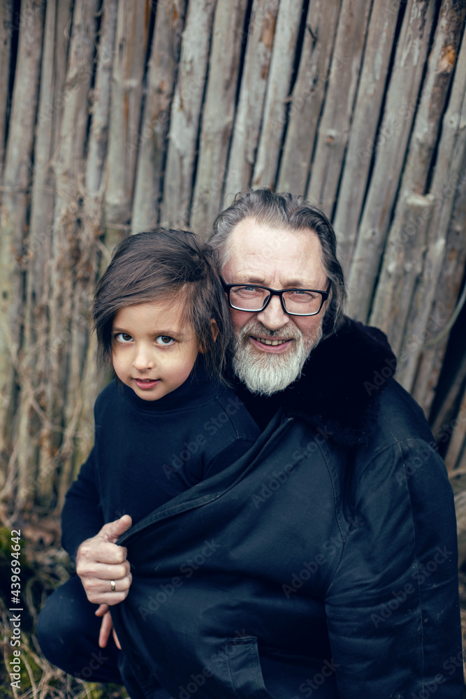 Fototapeta premium grandson in a blue hat and boots with a grandfather in a sheepskin coat stand at a wooden house shed in the village in spring in Russia