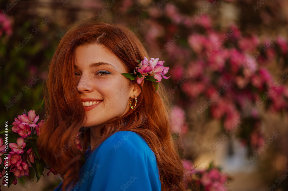 Fototapeta premium Happy smiling redhead freckled woman with blue eyeliner makeup posing near pink blooming tree. Copy, empty space for text