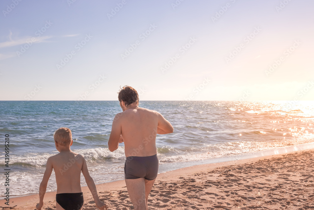 A man and a child walk along the sandy shore to the sea, illuminated by bright sunlight. Summer vacation and recreation, travel and tourism. Copy space