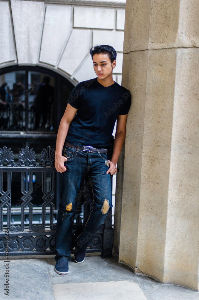 Dressing in a black T shirt and fashionable pants, leaning on a column and standing by a black metal railing, a young attractive asian teenager is looking down and thinking.