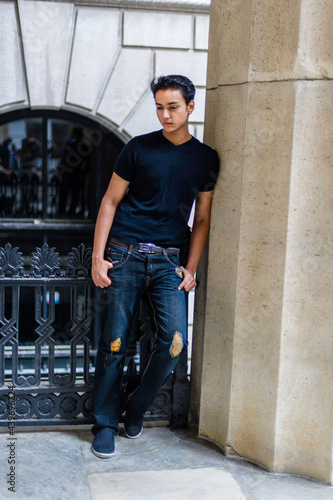 Obraz na płótnie Dressing in a black T shirt and fashionable pants, leaning on a column and standing by a black metal railing, a young attractive asian teenager is looking down and thinking. w salonie