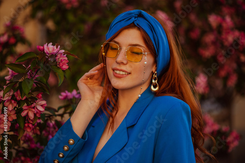Happy smiling redhead woman wearing trendy blue turban headband, yellow sunglasses with chain, stylish earrings, blazer. Spring, summer fashion, lifestyle conception. Copy, empty space for text