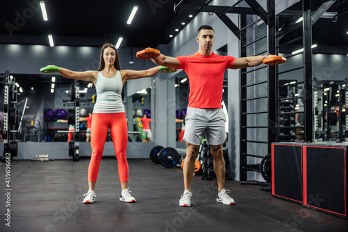 A muscular man and a beautiful young woman doing some arm and shoulder exercises together in the indoor gym at night time. Couple goals, fitness goals