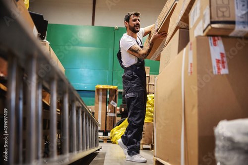 Hardworking tattooed bearded blue collar worker in overalls putting big heavy box on pile while standing in warehouse.
