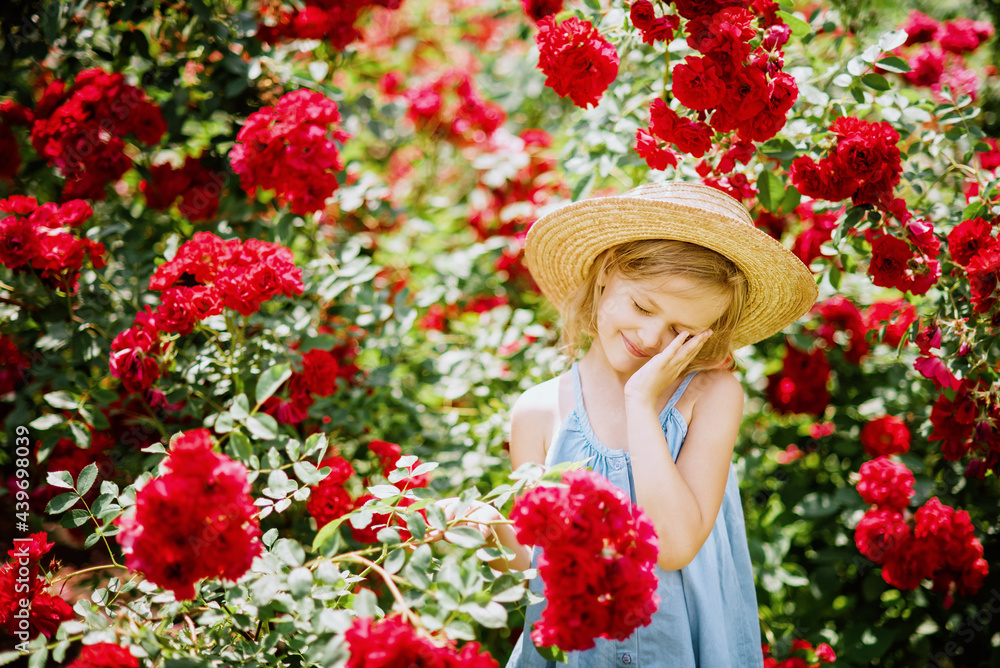 Fototapeta premium Summer portrait of pretty little girl wearing white dress and hat, posing in rose garden