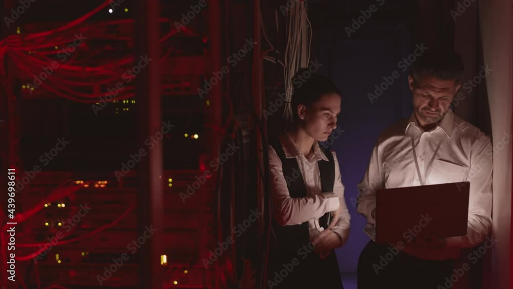 Medium shot of female and male Caucasian coworkers wearing formal clothing, using laptop computer in dark server room, talking Medium shot of female and male Caucasian coworkers wearing formal clothing, using laptop computer in dark server room, talking
