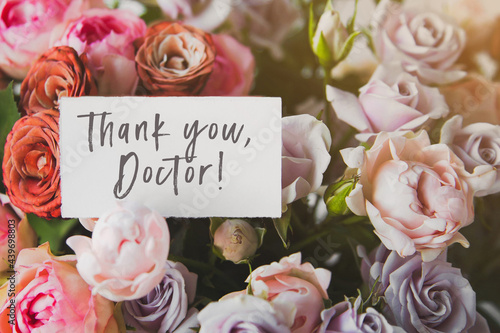 white gift card with the inscriptionThank you doctor in a bouquet of bright beautiful multi-colored roses, the concept of congratulations on the holiday 