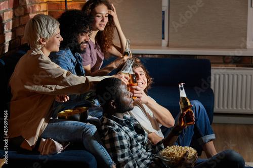 Multi-ethnic group of friends having small home party, meeting, sitting on couch, clinking bottles of beer, spending, enjoying time together, watching tv at night. side view