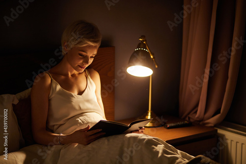 Beautiful female in pajamas lying on bed reading book, alone at night. Caucasian short haired lady in bedroom, charming cute woman in room lighted by lamp