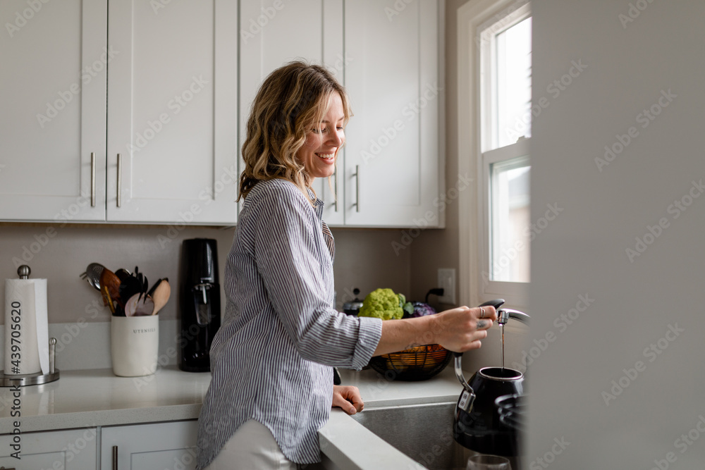 Smiling Woman Stands at Kitchen Sink and Fills a Teapot With Water Smiling Woman Stands at Kitchen Sink and Fills a Teapot With Water