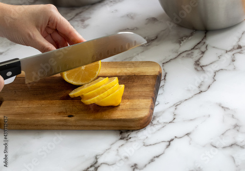 Petite planche à découper en bois sur une surface de marbre avec main qui tient un couteau en métal à lame droite et manche noir en train de couper un citron jaune en tranche dans la cuisine
