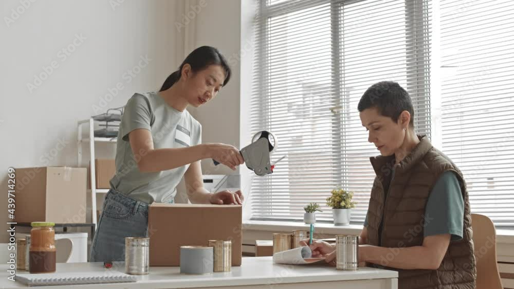 Medium slowmo of two female volunteer workers getting cardboard donation boxes for charity, filling them with eatables and sealing Medium slowmo of two female volunteer workers getting cardboard donation boxes for charity, filling them with eatables and sealing