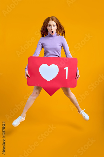 Shocked jumping woman with heart button, redhead female holding like icon of social media, emoji sign to follow subscribe interesting blog, popular forum. indoor studio shot isolated on yellow photo