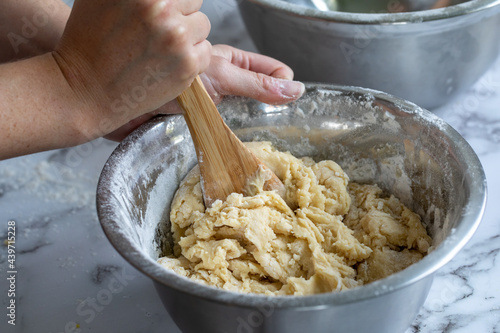 mains d'une femme en train de mélanger une pâte fraichement préparée avec des ingrédients simples dans un bol à mélanger rond en métal avec lumière naturelle et cuiller de bois