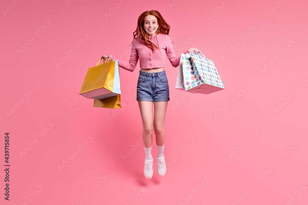 Happy trendy stylish carefree woman rejoicing with sales, shopping time, isolated on pink studio background, wearing casual outfit, denim jeans and pink shirt. portrait of jumping lady with red hair Happy trendy stylish carefree woman rejoicing with sales, shopping time, isolated on pink studio background, wearing casual outfit, denim jeans and pink shirt. portrait of jumping lady with red hair