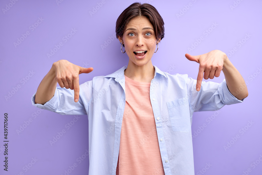Cheerful woman with short hair pointing index finger down, showing. copy space, free space for advertisement. Caucasian woman smiling at camera, isolated purple background Cheerful woman with short hair pointing index finger down, showing. copy space, free space for advertisement. Caucasian woman smiling at camera, isolated purple background