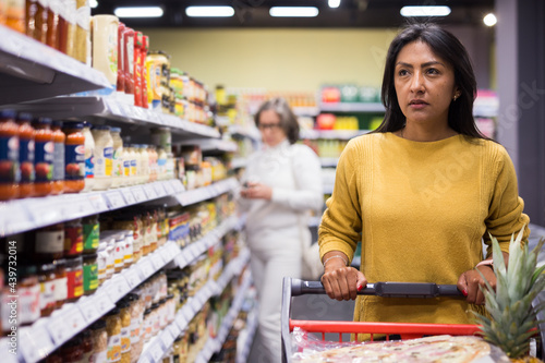 Portrait of thoughtful Hispanic woman walking with shopping trolley among shelves with products in grocery shop