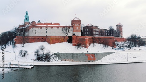 Drone view of the Wawel Royal castle. Vistula River bank in the foreground. Panoramic view of the city skyline during the winter season. Clear white sky in the background. Snow-Covered streets. 