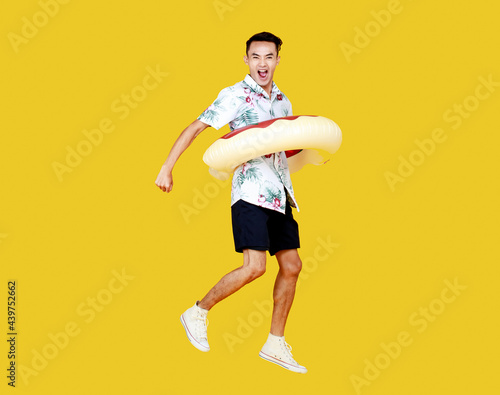 Young attractive Asian man wearing hat and white Hawaiian shirt and donut swim ring around his waist jumping with excitement against yellow background. Concept for beach vacation holiday