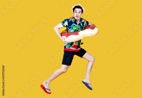 Young attractive Asian man wearing green and blue Hawaiian shirt and donut swim ring around his waist running against yellow background. Concept for holiday beach vacation