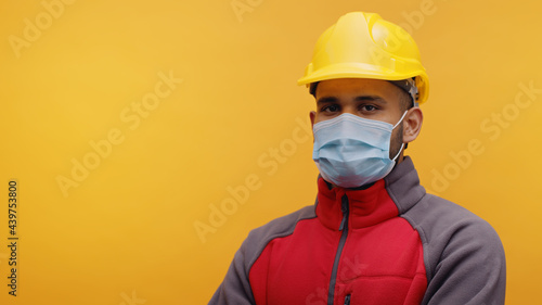 Portrait of a young Indian Engineer wearing safety yellow helmet and a mask standing with arms crossed in the studio with yellow background. Isolated man. Closeup shot.  © CameraCraft