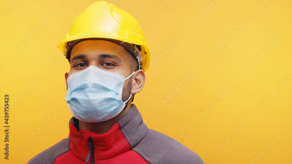 Portrait of a young Indian Engineer wearing safety yellow helmet and a mask standing in the studio with yellow background. Isolated man. Positive facial expression. Closeup shot. Portrait of a young Indian Engineer wearing safety yellow helmet and a mask standing in the studio with yellow background. Isolated man. Positive facial expression. Closeup shot.