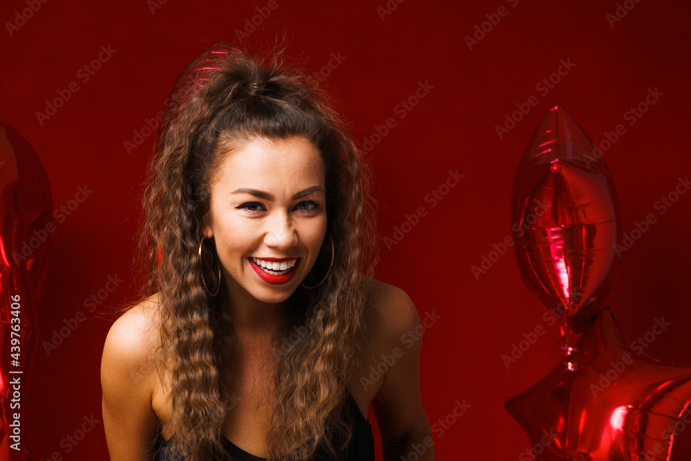 Portrait of a beautiful woman on a red background with red balloons posing in a black dress. The girl smiles with snow-white teeth on a holiday. Happy mood caucasian lady