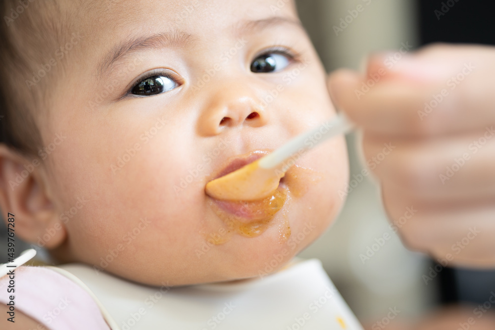 Happy infant baby sits in a child's chair eating first meal.Mom feeds baby. Happy infant baby sits in a child's chair eating first meal.Mom feeds baby.